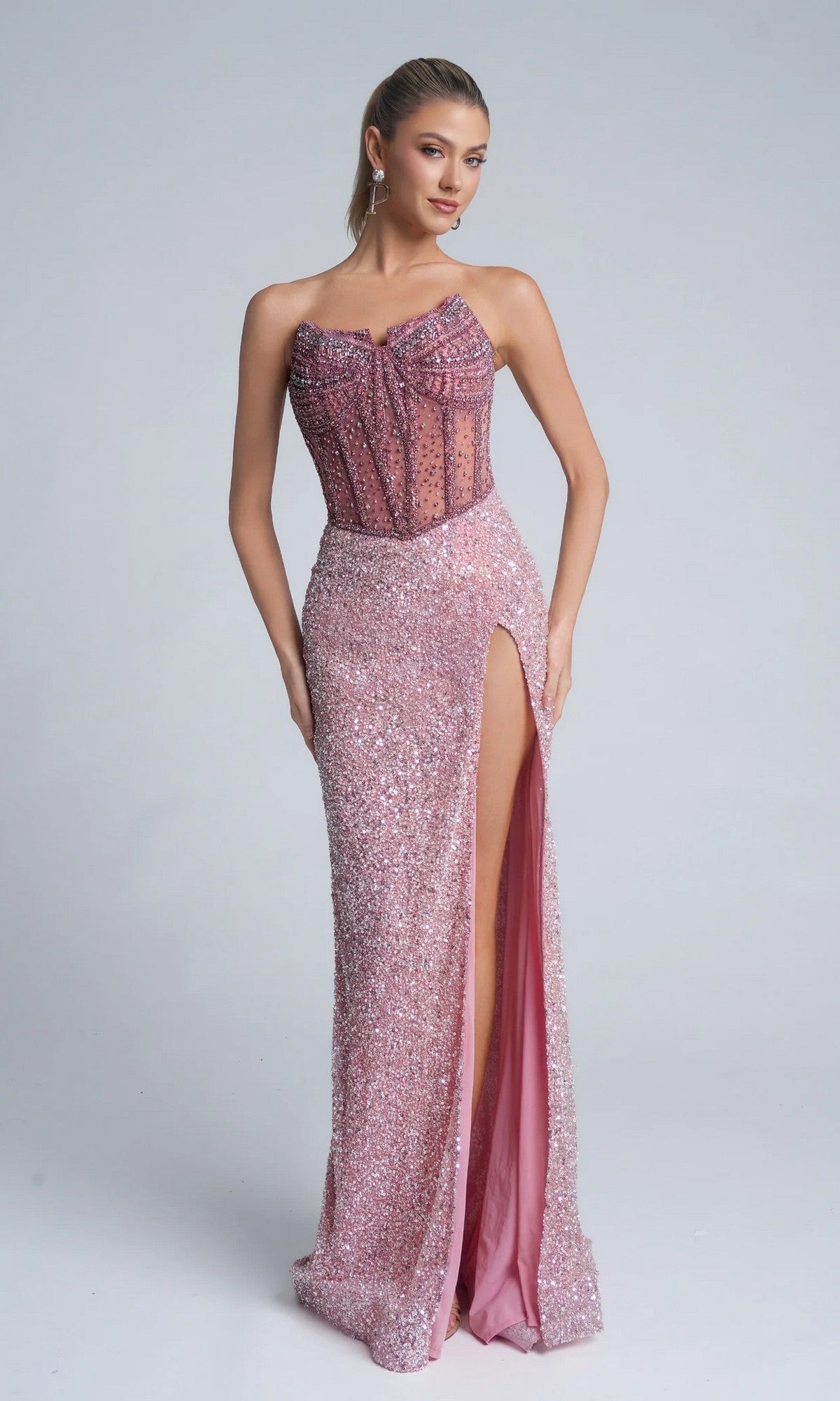 A woman poses against a plain light background wearing the Portia and Scarlett PS25990C long pink prom dress, featuring a strapless sparkly design, sheer corset bodice, and high leg slit.